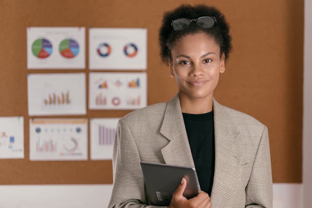 Professional woman holding a tablet in front of a presentation board displaying various market analysis graphs and charts related to real estate trends.