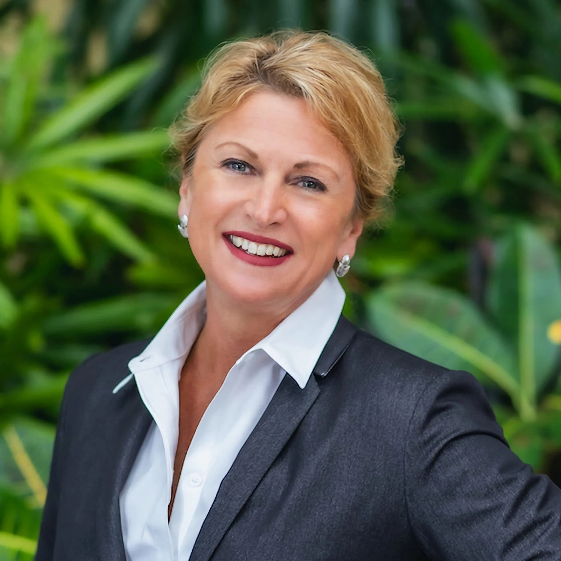 Maria Wilkes, luxury real estate advisor, smiling in a professional outfit against lush greenery, representing excellence in personalized real estate services.